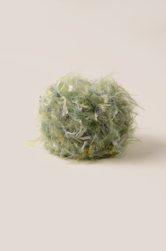 Lush Fern hand-blended yarn – muted grass green base with blue, yellow, and white threads, soft fluffy texture inspired by rainy meadows
